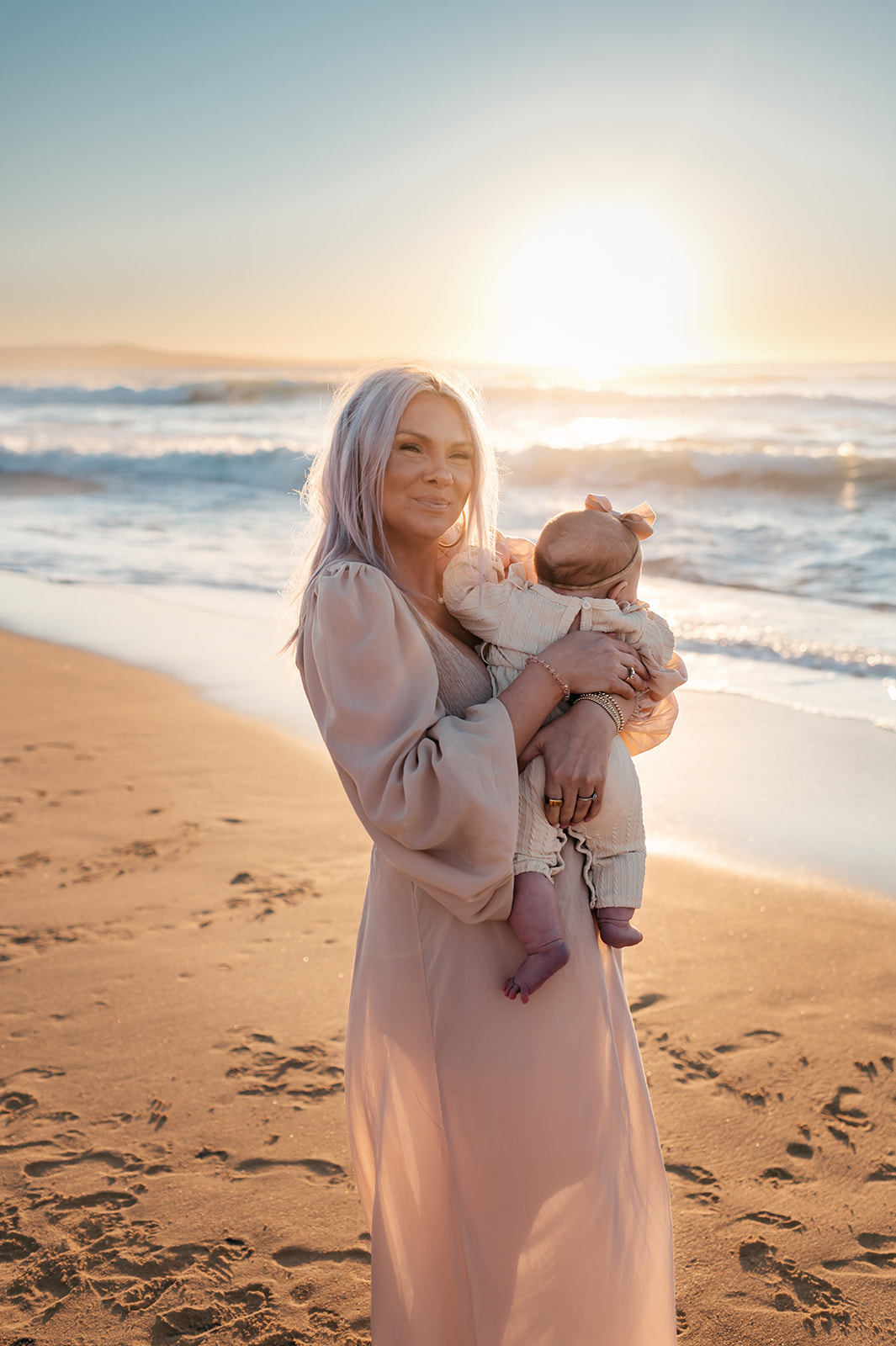 Mother in a flowy blush dress holding her baby at the beach during golden hour