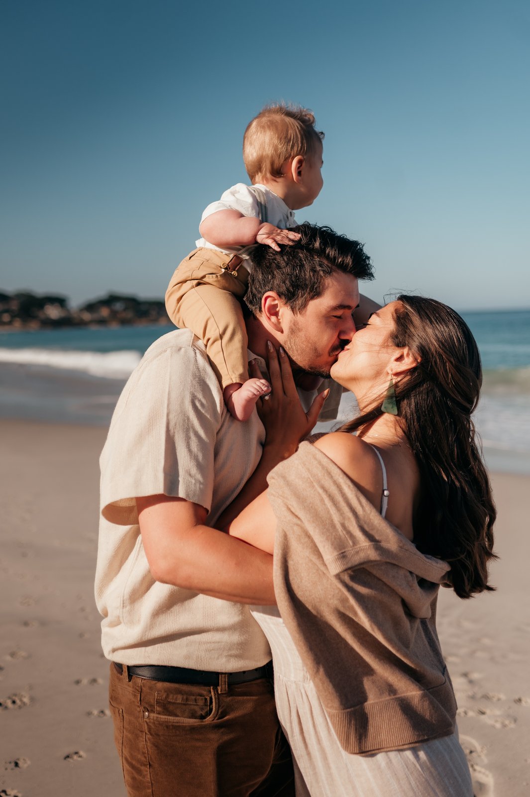 Mom and dad kissing on Carmel Beach while baby rides on dad's shoulders