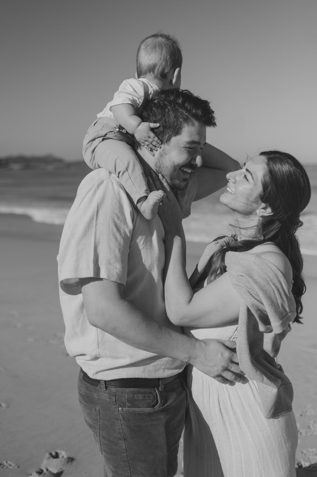 Black and white photo of parents laughing face to face with baby on dad's shoulders at Carmel Beach