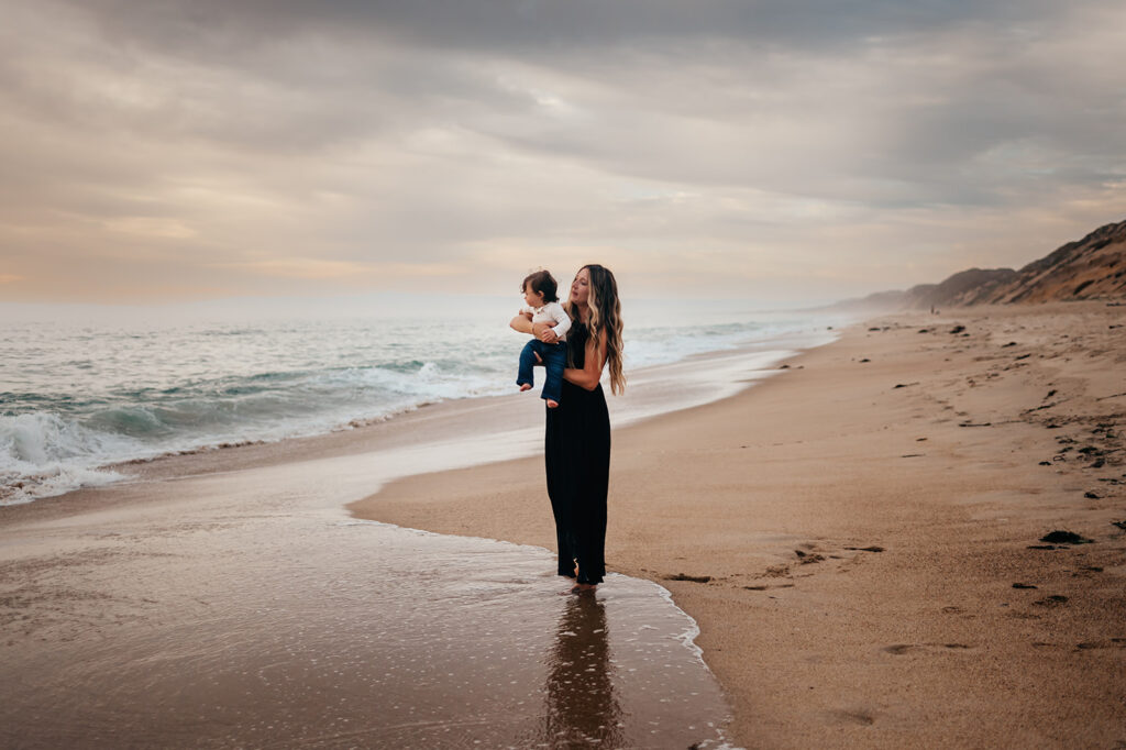 Mom standing in the surf holding her son during a motherhood session at Fort Ord Dunes State Park