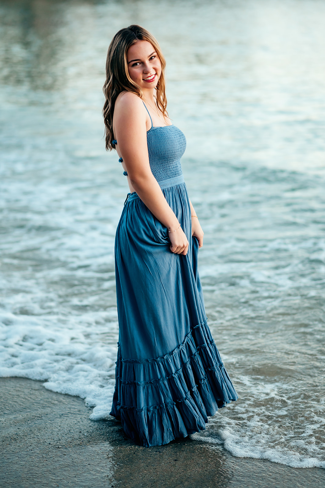 Senior in a long blue dress holding her skirt in the surf on the Monterey coast