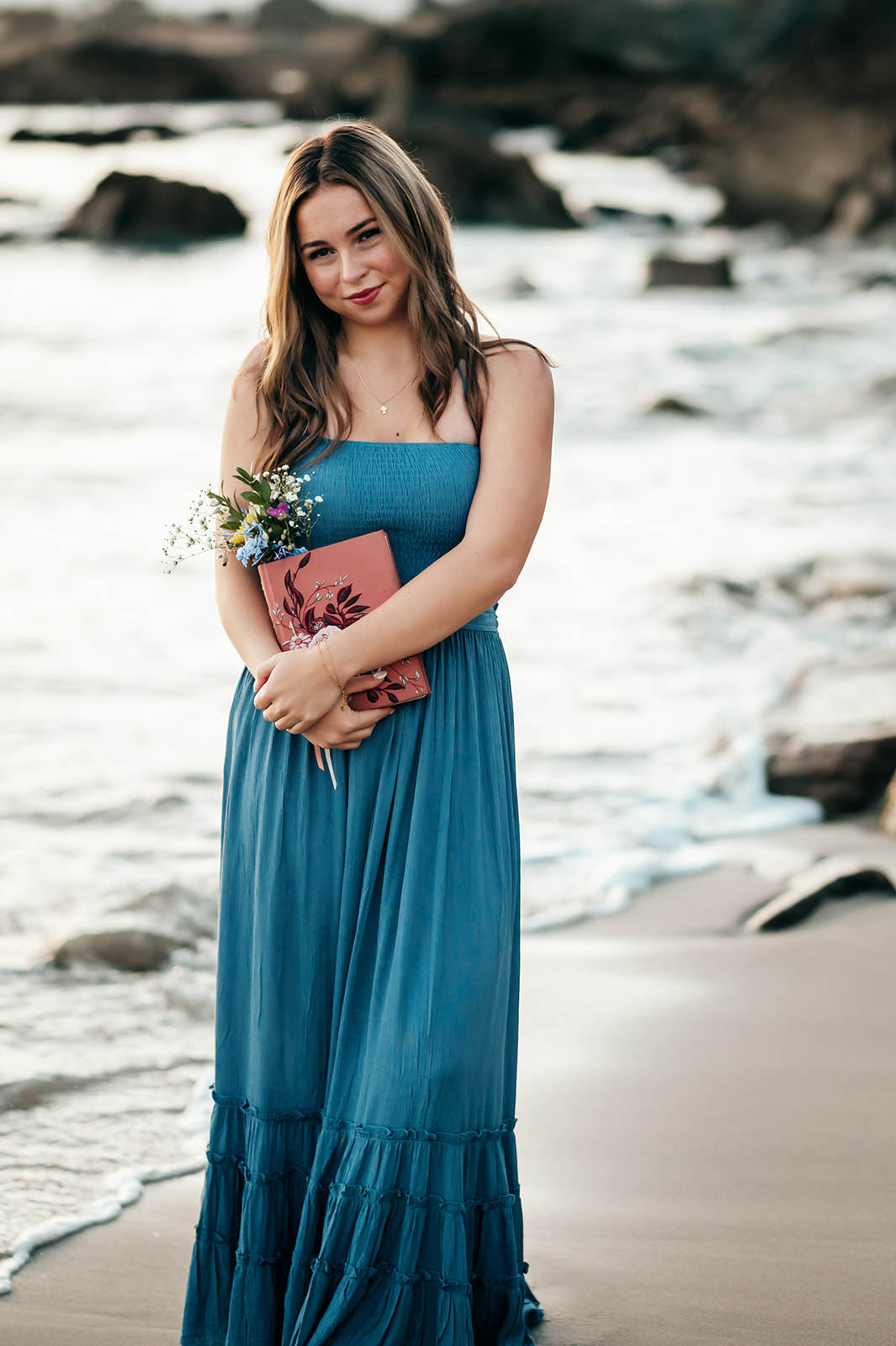 Full-length senior portrait in a blue maxi dress holding wildflowers on the beach