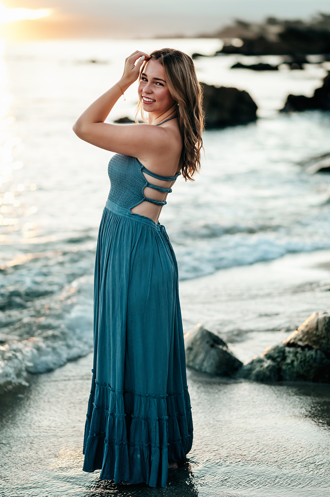 Senior girl in a flowy blue dress standing in the surf with sunrise flare at Lovers Point Beach