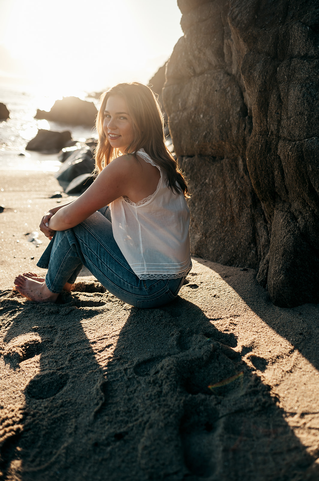 Relaxed senior portrait in jeans and a white top sitting barefoot in the sand at sunrise