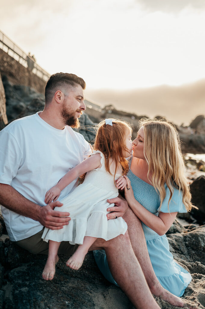Daughter kissing mom while sitting on her dad's lap at the beach in Pacific Grove, CA. 