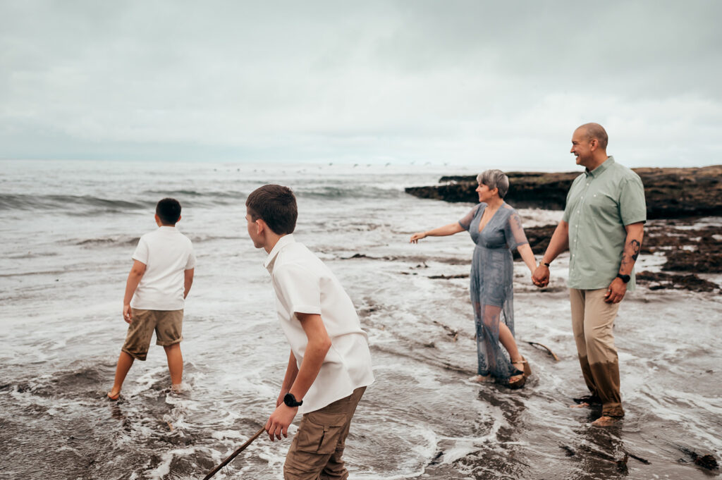 Parents holding hands in the shallow surf while their two sons play nearby during a Santa Cruz family session