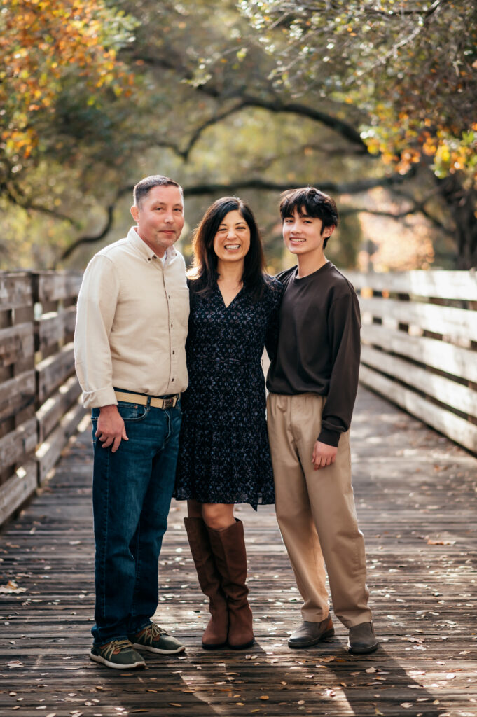 Family of three standing on a wooden bridge under oak trees turning gold, dressed in fall layers
