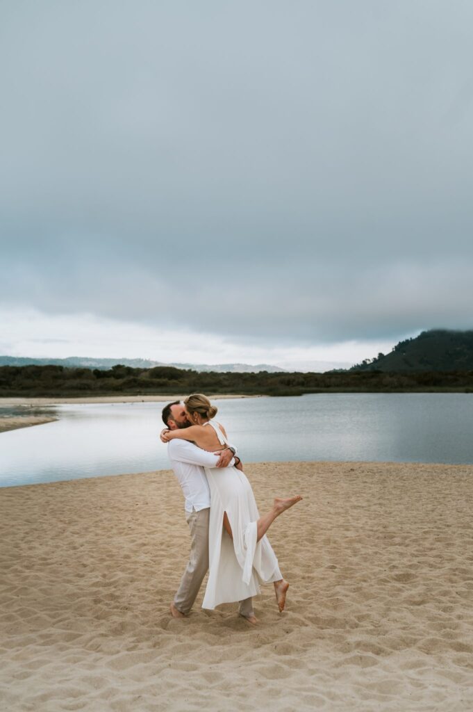 Husband lifting his wife off the sand in front of the Carmel River lagoon during a sunset family session