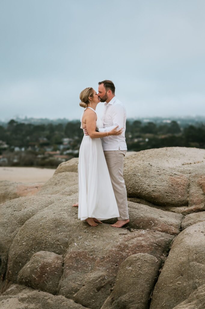 Couple kissing barefoot on the rocks above Carmel River Beach with Carmel-by-the-Sea behind them