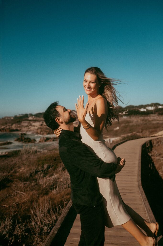 Joe lifting Peyton off her feet on the Asilomar boardwalk while she shows off her ring and laughs
