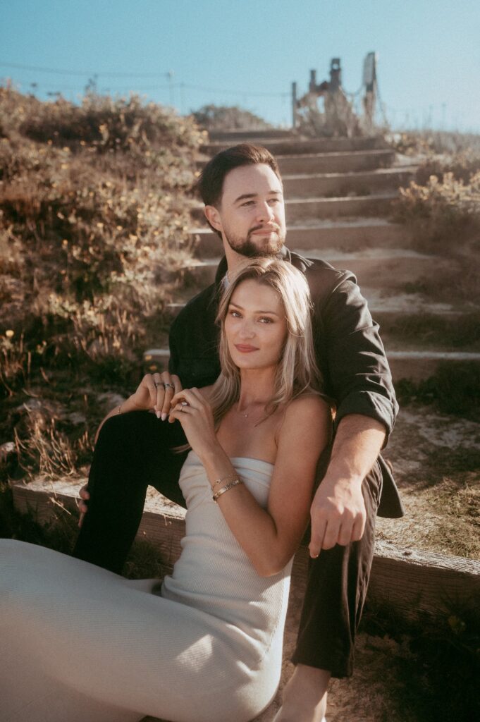 Joe and Peyton sitting together on the wooden steps in the Asilomar dunes at golden hour, Peyton showing her ring