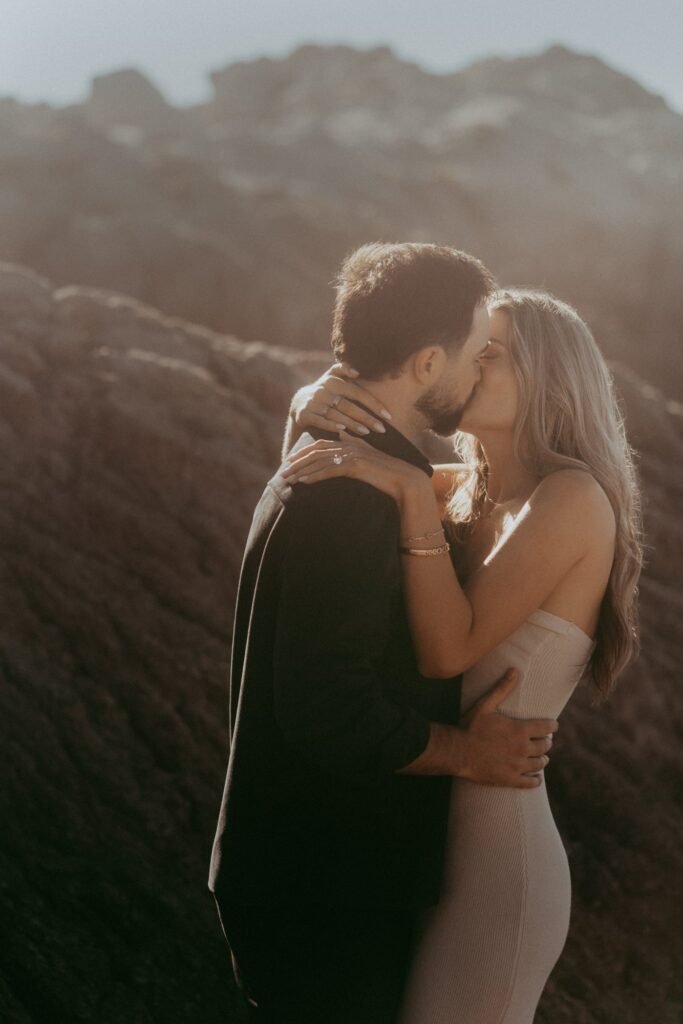 Joe and Peyton kissing in hazy backlit golden hour light with the Asilomar rocks glowing behind them