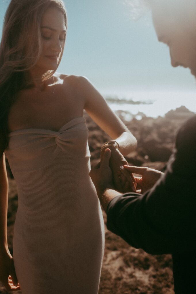 Joe sliding the engagement ring onto Peyton's finger, backlit by the sun with the ocean behind them at Asilomar