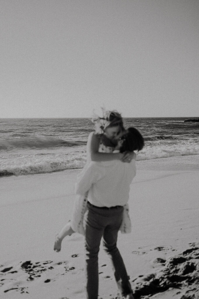 Black and white photo of Nick lifting Patrycja off her feet for a kiss on Garrapata Beach
