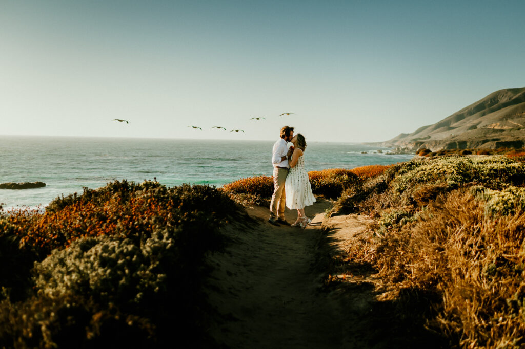 Nick and Patrycja kissing on the coastal trail at Garrapata State Park with pelicans flying overhead and the Big Sur mountains behind them