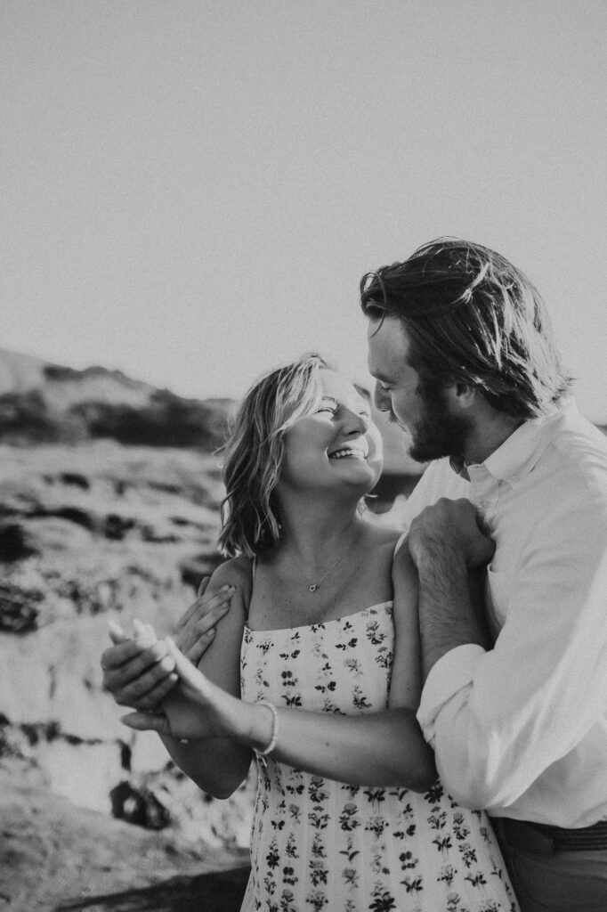 Black and white photo of Patrycja laughing up at Nick as he wraps his arms around her at Garrapata State Park