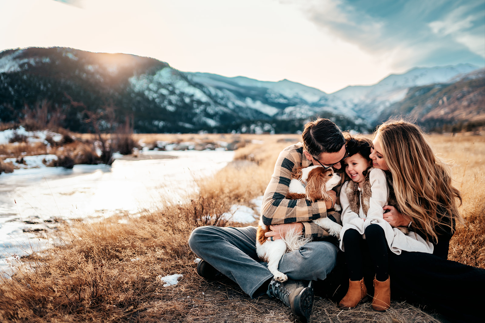 Parents and their young daughter snuggling their dog beside a snow-lined mountain river at sunset, family photography by Britt Honey Photography