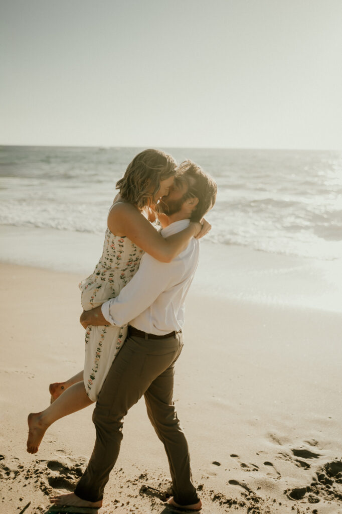 Nick holding Patrycja up for a kiss, barefoot on the sand at Garrapata Beach at golden hour