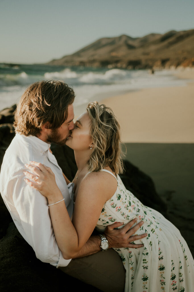 Nick and Patrycja sitting on the rocks kissing at Garrapata Beach with the Big Sur mountains behind them
