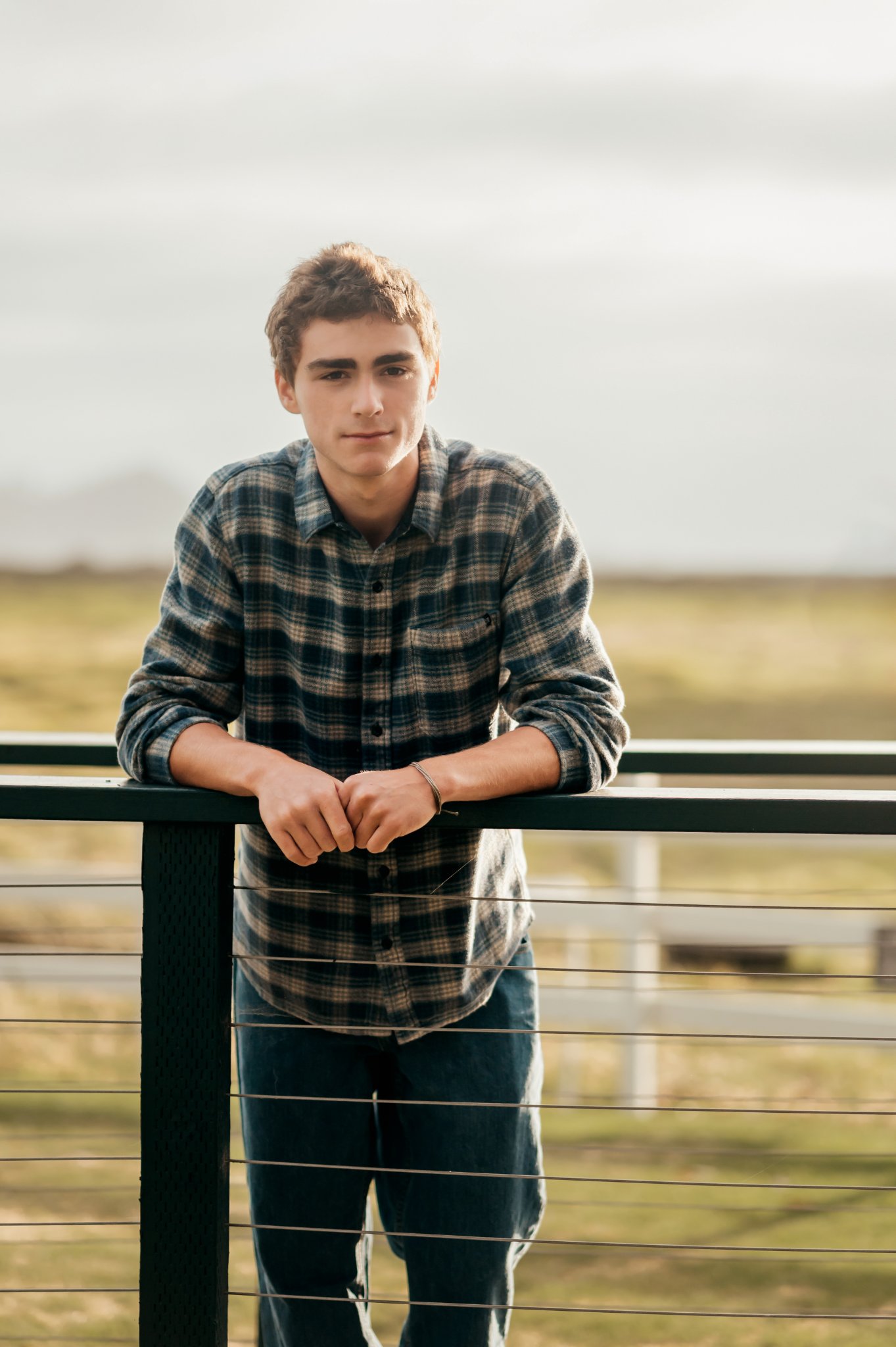 High school senior leaning on a dark bridge railing with the meadow behind him at Mission Ranch in Carmel
