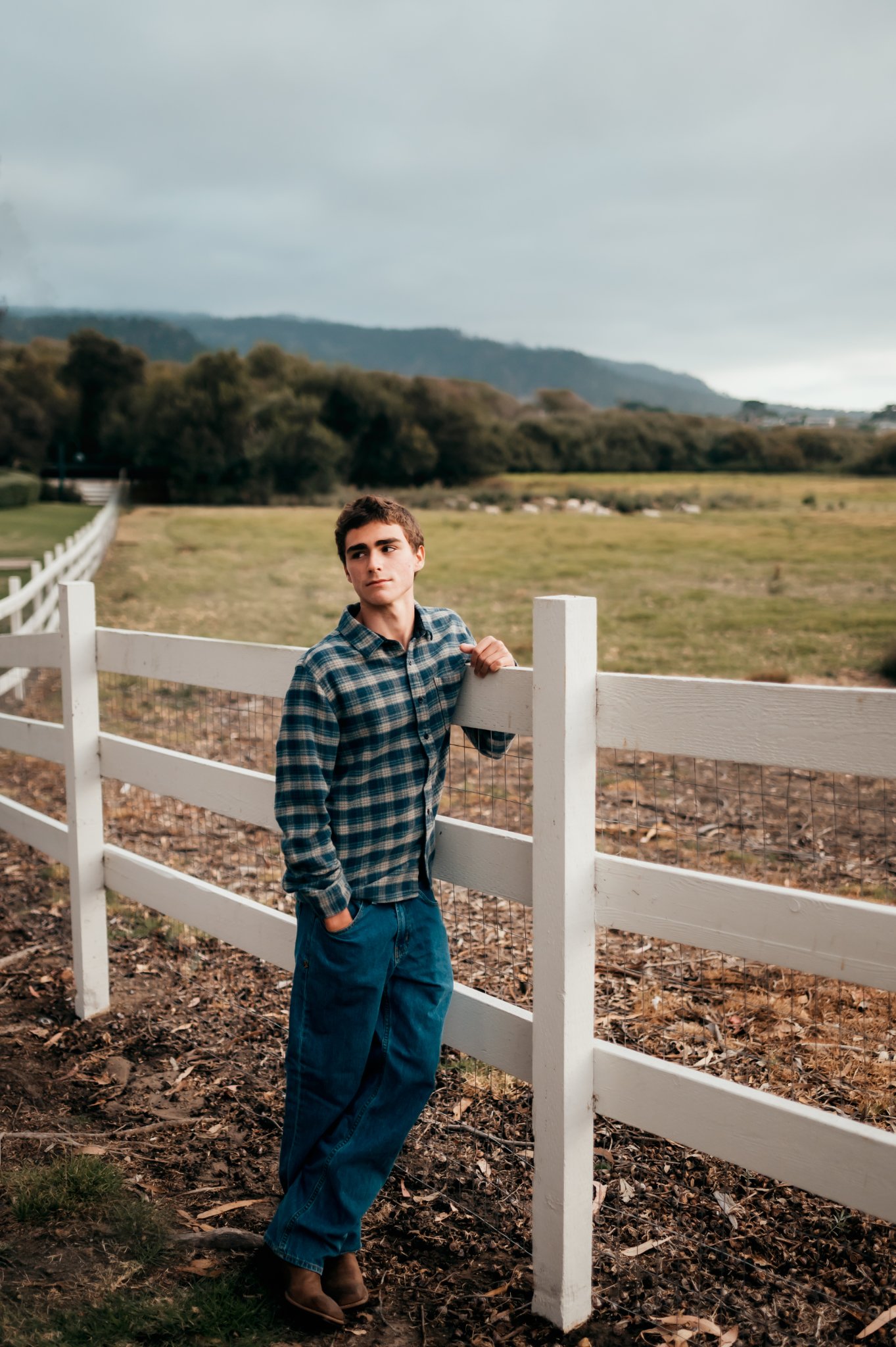 Senior guy in flannel and boots leaning on a white fence with the meadow and hills behind him, Carmel senior photos
