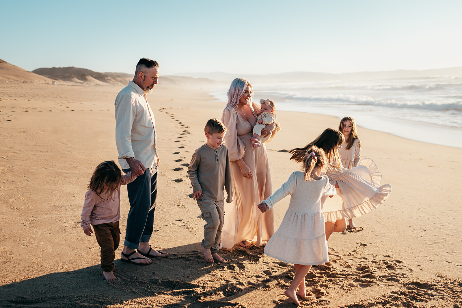 Family of seven walking on the sand at golden hour, mom holding the baby while the kids twirl, Monterey area beach family session by Britt Honey Photography