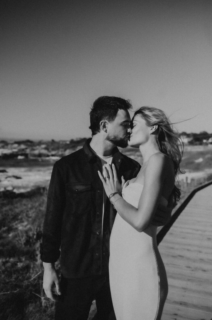 Black and white photo of Joe and Peyton kissing on the Asilomar boardwalk with her hair blowing in the wind