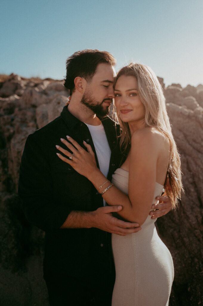 Joe holding Peyton close against the rocks at Asilomar in golden hour light, her engagement ring resting on his chest