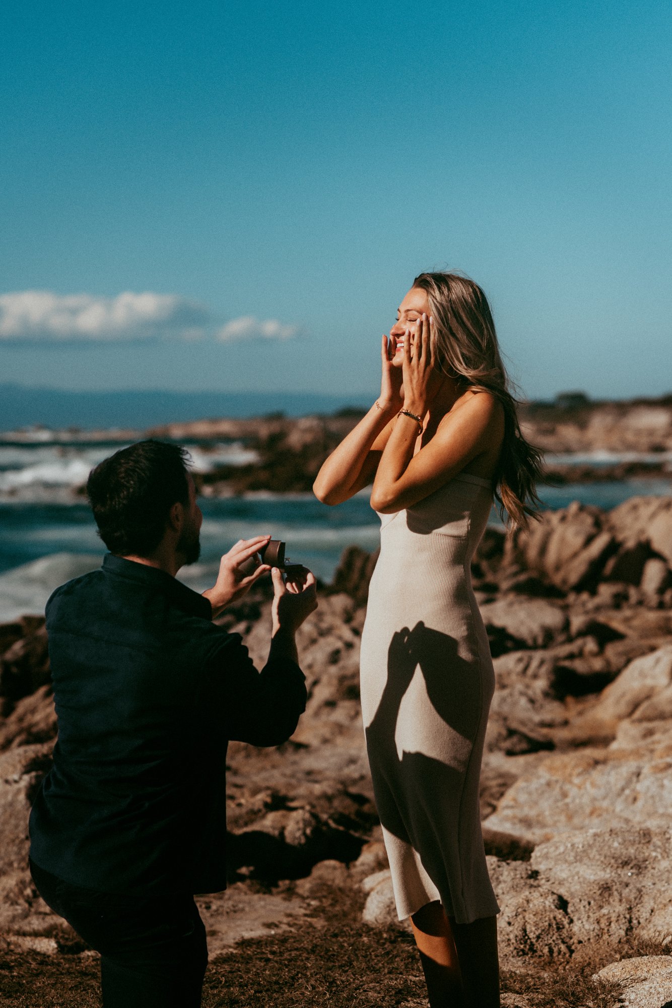 Joe on one knee holding the ring box open while Peyton covers her face and laughs on the rocks at Asilomar in Pacific Grove